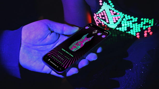 Smartphone displaying a JASON BRICKHILL garment’s digital identity beside an illuminated fashion piece.